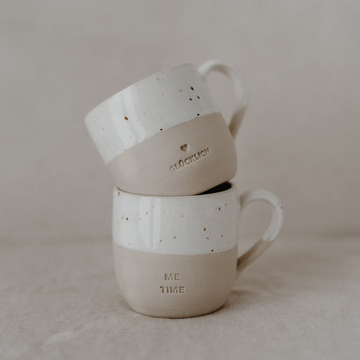 Eine beige-weiße Keramiktasse liegt auf der Seite auf einem Leinentuch. Die obere Hälfte ist glasiert und weiß mit kleinen braunen Sprenkeln, die untere Hälfte matt beige. Ein kleines, eingraviertes Herz ist an der Seite zu sehen. Auf dem Tassenboden steht in einem Kreis: ‚Zuhause bei mir‘.