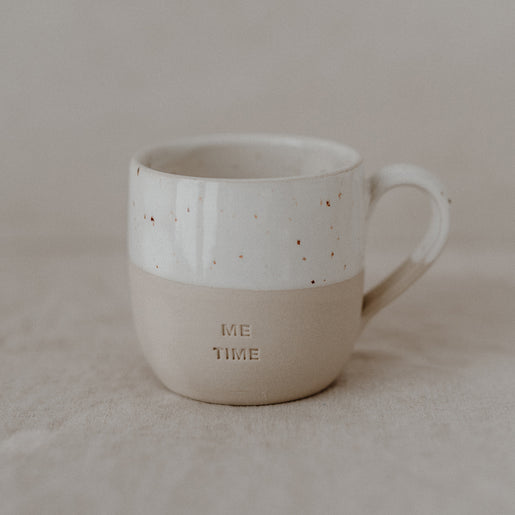 Eine cremefarbene Keramiktasse mit glatter Form, deren obere Hälfte weiß glasiert und braun gesprenkelt ist. Der untere, matte Teil ist beige und trägt die Prägung ‚ME TIME‘ in Großbuchstaben. Die Tasse steht auf hellem Leinenstoff vor neutralem Hintergrund.