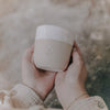 Zwei Hände halten einen minimalistischen Keramikbecher in sanftem Beige mit cremeweißer, gesprenkelter Glasur am oberen Rand. Auf der Vorderseite ist ein kleines Herz und darunter das Wort ‚TANTE‘ eingeprägt. Der Becher wird leicht schräg vor einem natürlichen, rauen Hintergrund präsentiert – ein stilvolles Geschenk mit persönlicher Botschaft.