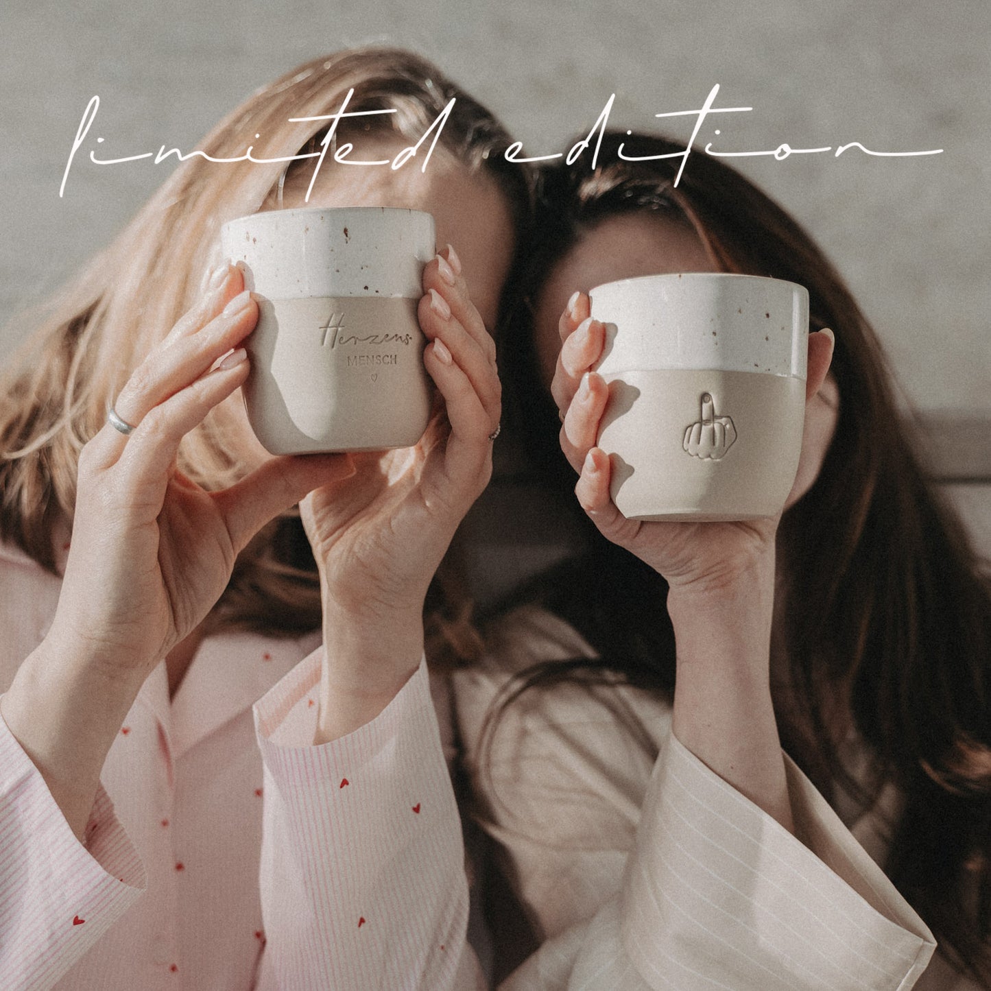 Zwei Frauen halten Herzensmensch- und Mittelfinger-Becher der Limited Edition vor sich, beige-weiß mit Keramikstruktur.