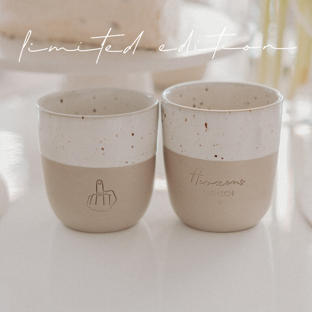Zwei Limited-Edition-Becher in Beige und Weiß mit Herzensmensch- und Mittelfinger-Motiv auf hellem Tisch arrangiert.