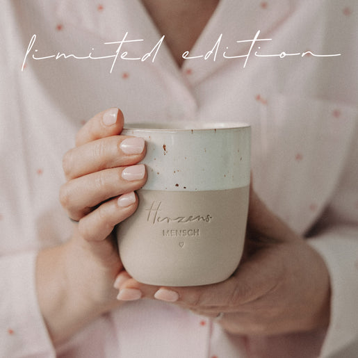 Person hält beige-weißen Limited-Edition-Becher mit Herzensmensch-Gravur nah vor dem Körper in gemütlicher Atmosphäre.