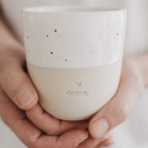 Keramikbecher „Onkel“ mit Herzprägung wird in beiden Händen gehalten, beige und weiß gesprenkelte Optik.
