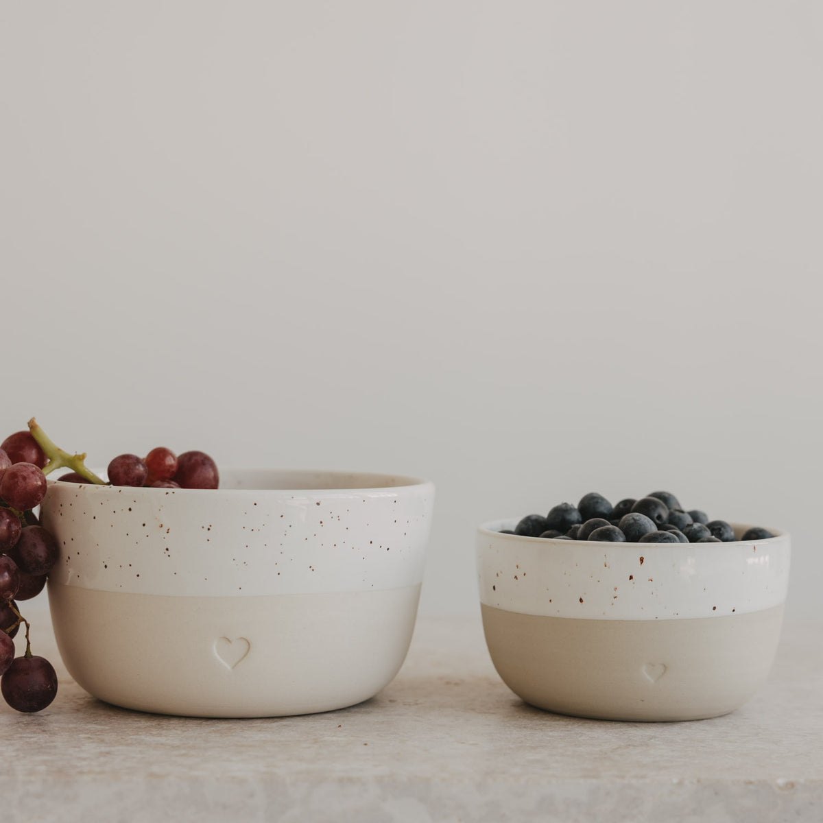 Große und kleine Keramikschale mit Herz in Beige und Weiß, gefüllt mit Trauben und Heidelbeeren