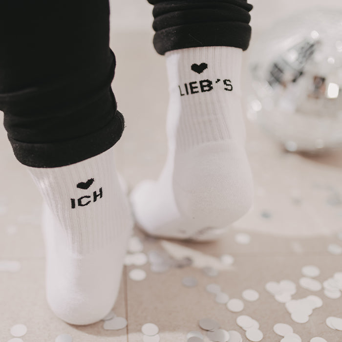 Weiße Socken mit schwarzem Herz und den Schriftzügen „ICH“ und „LIEB’S“, getragen zu schwarzer Hose und umgeben von silbernem Konfetti.