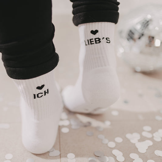 Weiße Socken mit schwarzem Herz und den Schriftzügen „ICH“ und „LIEB’S“, getragen zu schwarzer Hose und umgeben von silbernem Konfetti.