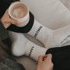 Weiße Socken „Erstmal Kaffee“ beim Entspannen mit Kaffeetasse auf Sofa