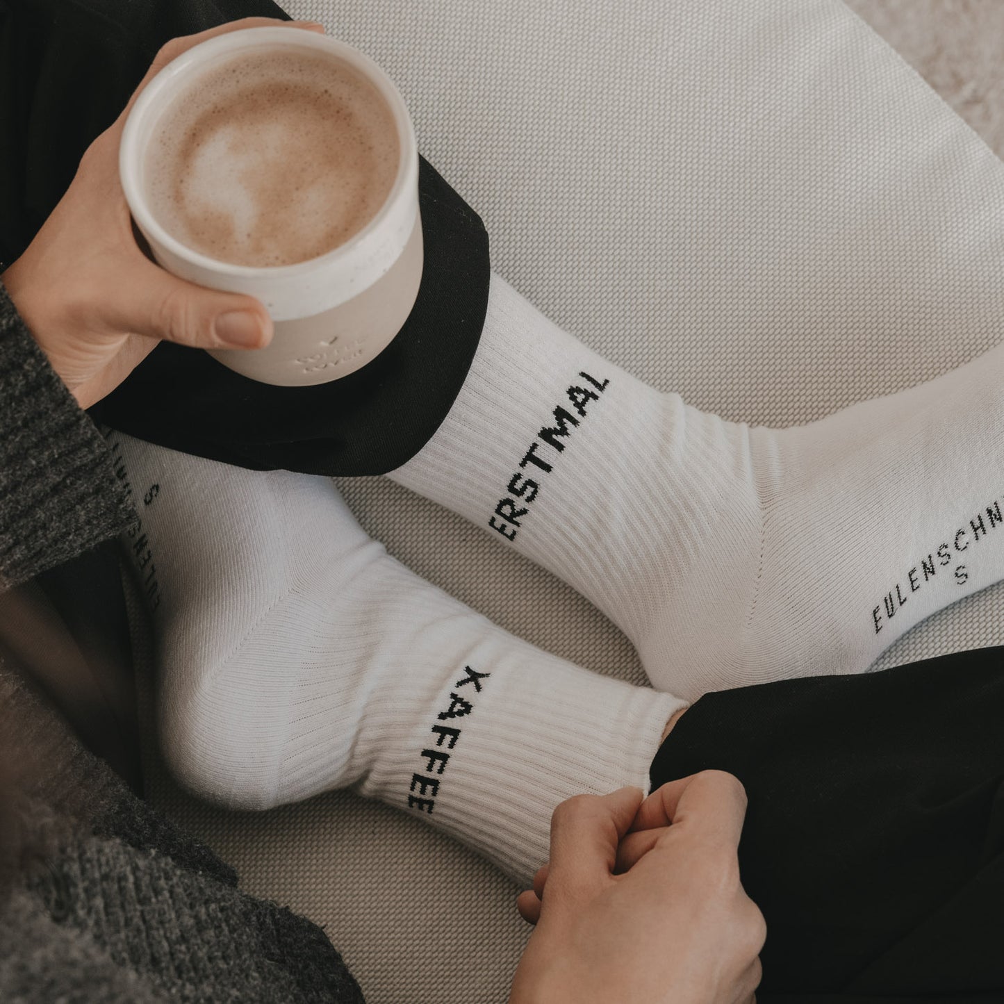 Weiße Socken „Erstmal Kaffee“ beim Entspannen mit Kaffeetasse auf Sofa
