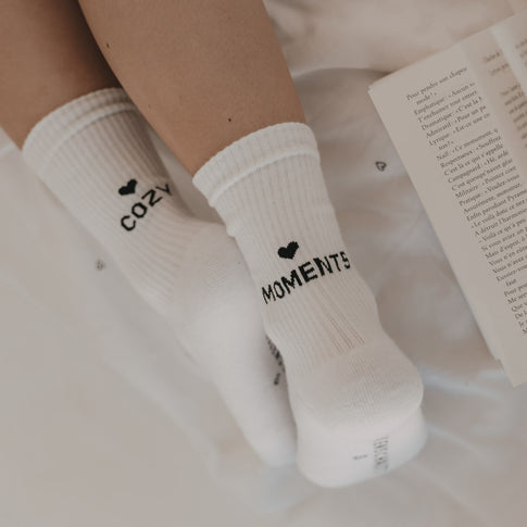 Weiße Socken „Cozy Moments“ in gemütlicher Atmosphäre mit Buch und Textil im Hintergrund