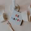 BE HAPPY Streichholzschachtel neben Kerze und Stoffband, dekorativ arrangiert.