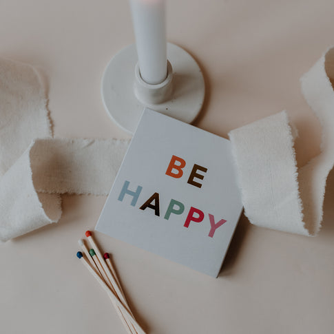BE HAPPY Streichholzschachtel neben Kerze und Stoffband, dekorativ arrangiert.