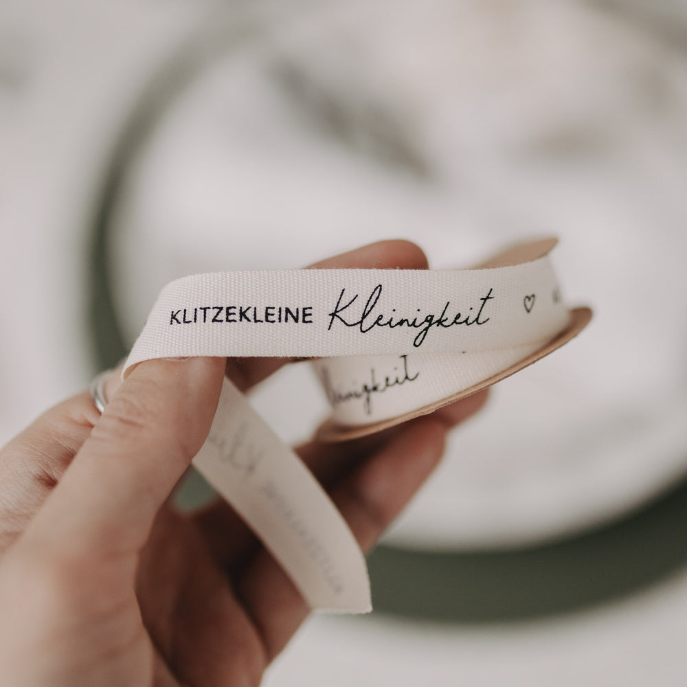 Hand hält weißes Stoff-Geschenkband mit Aufdruck „Klitzekleine Kleinigkeit“.