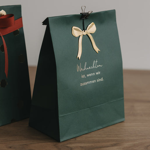 Grüne Papiergeschenktüte mit goldener Schleife und Aufdruck „Weihnachten ist, wenn wir zusammen sind"