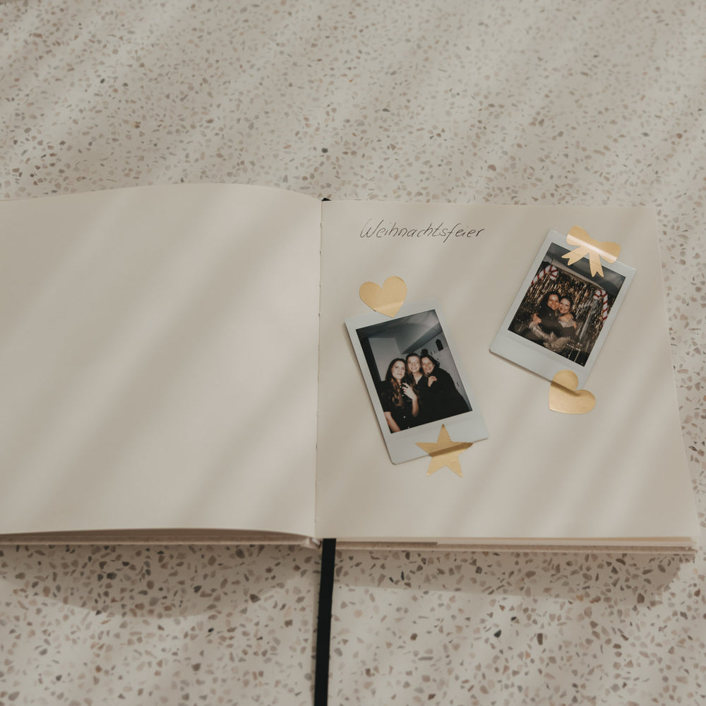 Offenes Buch mit Blankoseiten, eingeklebten Fotos und goldenen Herz-, Stern- und Schleifenstickern