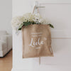 Jutetasche mit weißem Schriftzug „Hier steckt ganz viel Liebe drin“, gefüllt mit weißen Hortensien und gepunktetem Tuch, hängt an einer weißen Tür – liebevoll dekoriert.