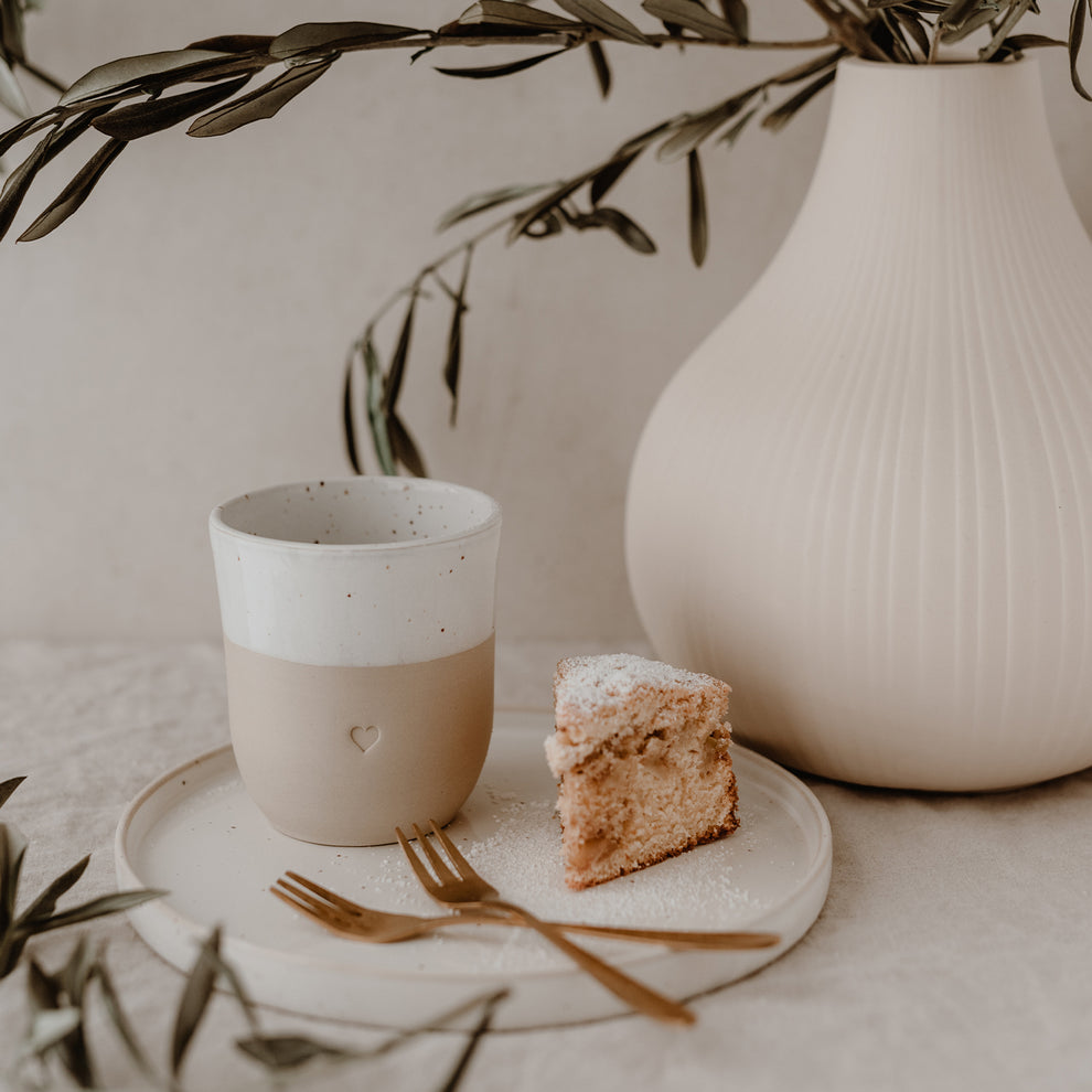 Keramikbecher mit Herz und Kuchenteller auf hell gedecktem Tisch, daneben Vase mit Olivenzweigen – sanftes, stilvolles Setting.